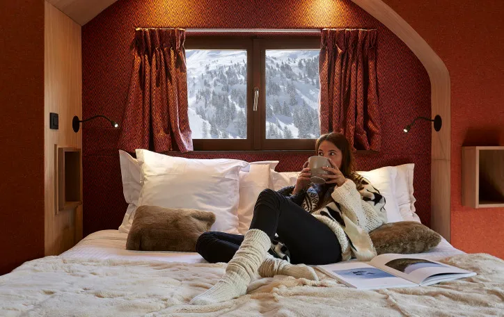 A person drinks a hot chocolate on the bed of the room of the Hotel des Dromonts